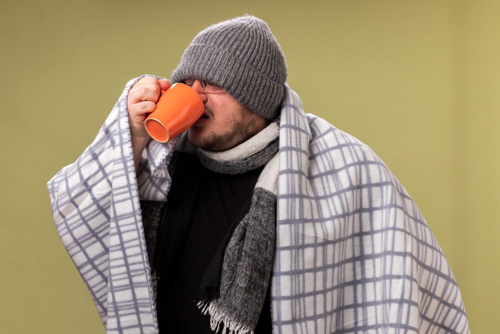 weak middle aged ill male wearing winter hat scarf wrapped plaid drinks tea from cup isolated olive green wall