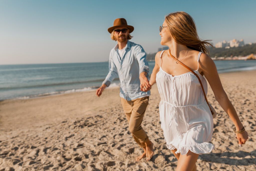 Smiling man hat blond woman white dress running together beach